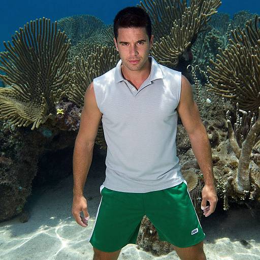 Photograph of a muscular, dark-haired man in a white sleeveless polo and green shorts, standing underwater with coral reefs behind him.