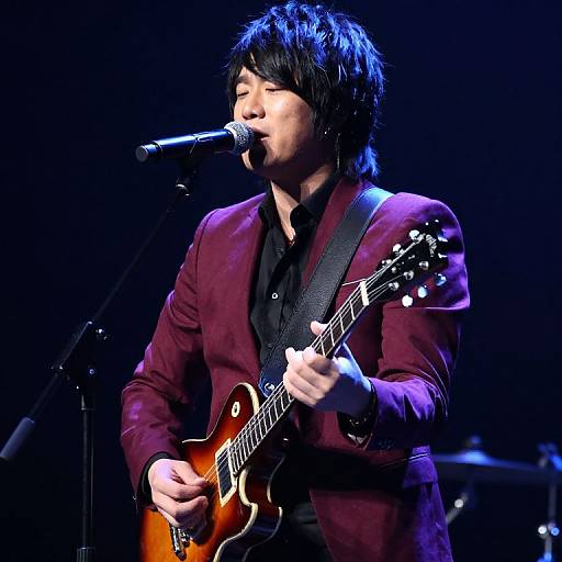 Photograph of an Asian male musician with black hair, wearing a maroon blazer and black shirt, singing into a microphone while playing a sunburst