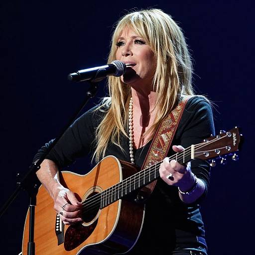 Photograph of a blonde woman with long hair, wearing a black top, singing into a microphone while playing an acoustic guitar on stage.