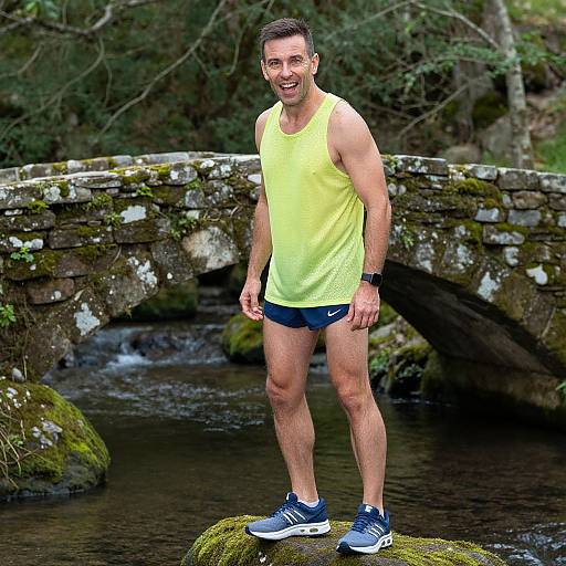 Photograph of a smiling, fit man in a yellow tank top and blue shorts, standing on a mossy rock by a mossy stone bridge over
