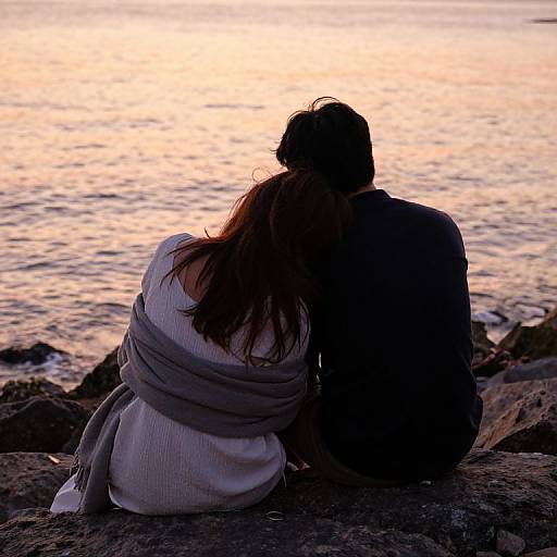 Silhouetted couple sitting on rocky shore, back to camera, wrapped in blankets, watching sunset over calm water. Romantic, peaceful evening scene.