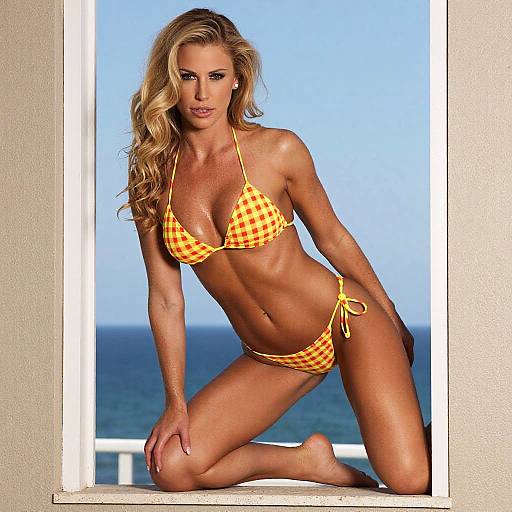 Photograph of a blonde woman with wavy hair, wearing a yellow-orange checkered bikini, kneeling on a balcony overlooking the ocean.