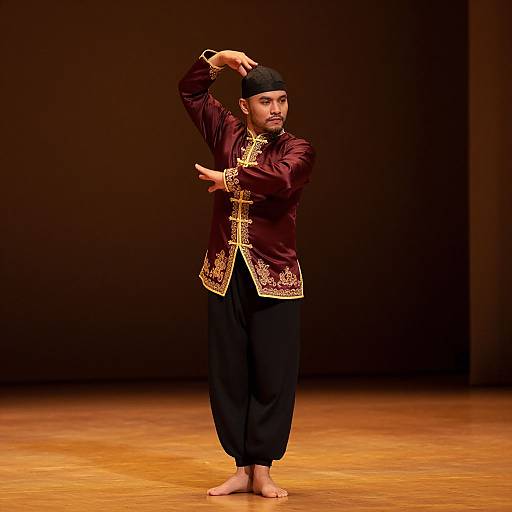 Photograph of a South Asian man performing traditional dance. He wears a maroon and gold embroidered tunic, black pants, and a black cap,