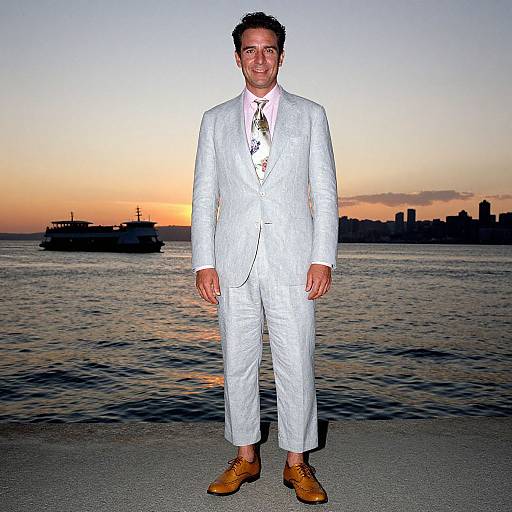 Photograph of a smiling man in a white pinstripe suit, brown shoes, and patterned tie, standing on a waterfront at sunset with a