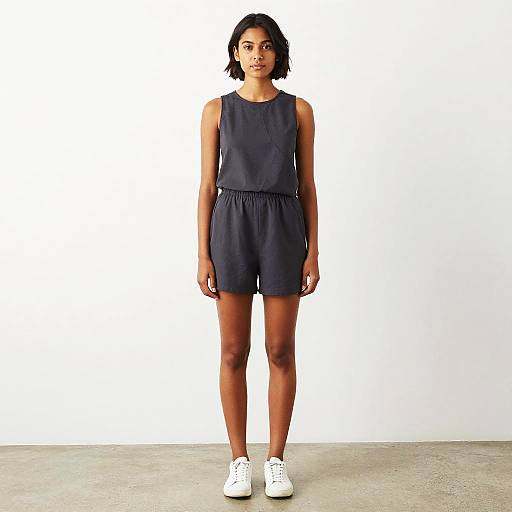 Photograph of a young woman with short black hair, wearing a dark navy sleeveless romper and white sneakers, standing against a plain white background.