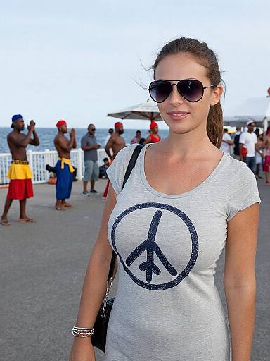 Photograph of a smiling woman with brown hair in a ponytail, wearing sunglasses, white peace sign t-shirt, standing at a beachfront, surrounded
