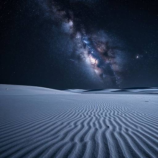 Photograph of a starry night sky above a rippled, snow-covered desert landscape. Milky Way galaxy visible, with bright stars and galaxies stretching across