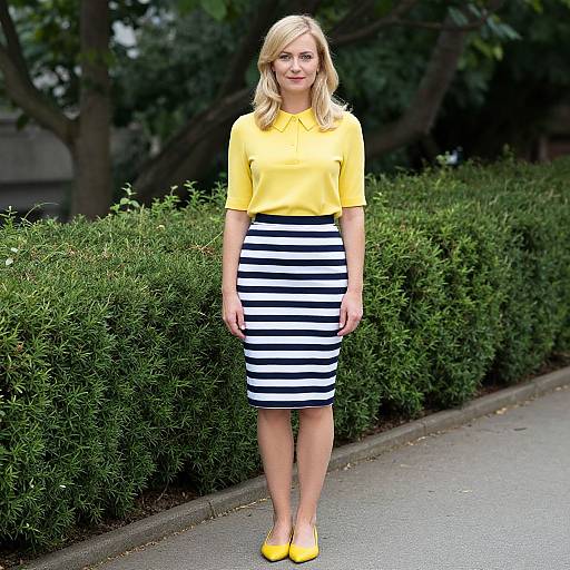 Photograph of a blonde woman in a yellow polo shirt and black-and-white striped skirt, standing on a sidewalk with green hedges in the background,