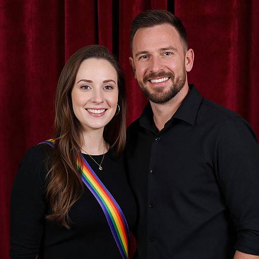 Photograph of a smiling couple; woman with long brown hair, rainbow sash, black top; man with short brown hair, beard, black shirt