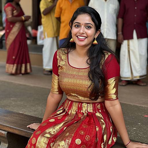 Photograph of a smiling Indian woman with long black hair, wearing a gold and red traditional saree, seated on a wooden bench, with people in