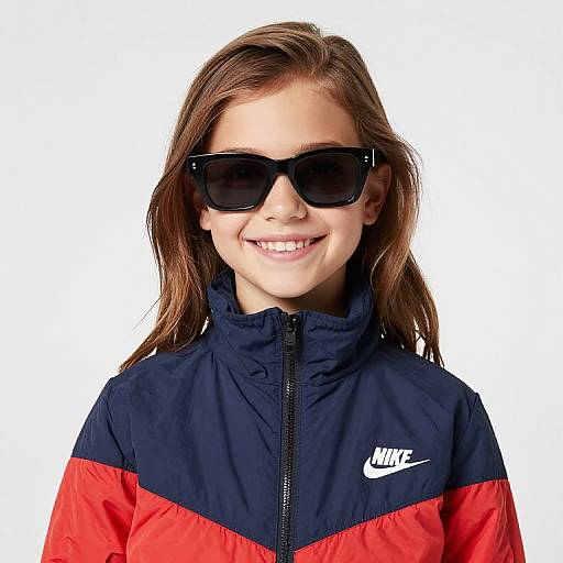 Photograph of a smiling young girl with light brown hair, wearing black sunglasses, a navy and red Nike sports jacket, and white background.