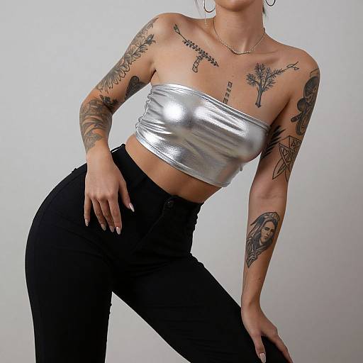 Photograph of a tattooed woman with light skin, wearing a silver metallic tube top and black high-waisted pants, posing against a plain white