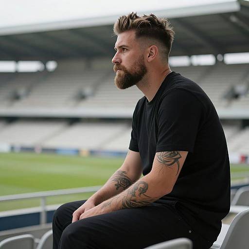 Photograph of a bearded, tattooed man with spiked hair, wearing a black t-shirt and black pants, sitting in a stadium.