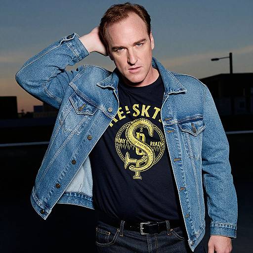 Photograph of a white man with short brown hair, wearing a blue denim jacket, black shirt with yellow snake emblem, black jeans, and black belt