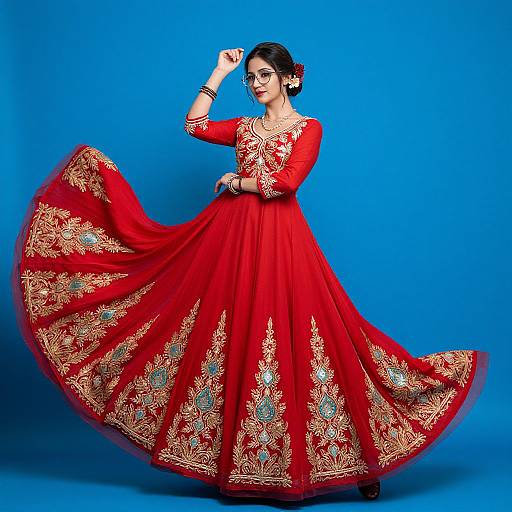 Photograph of an Indian woman in a vibrant red, full-length lehenga with gold and blue embroidery, striking a pose against a solid blue background.