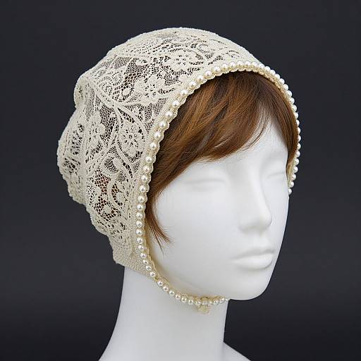 Photograph of a white mannequin head wearing an intricate, cream-colored lace bonnet adorned with pearl beads along the edge.
