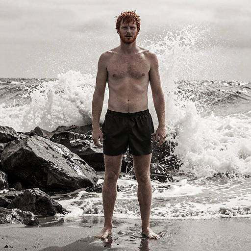 Photograph of a shirtless, red-haired, bearded man in black swim trunks standing on a rocky beach with large, crashing waves behind him