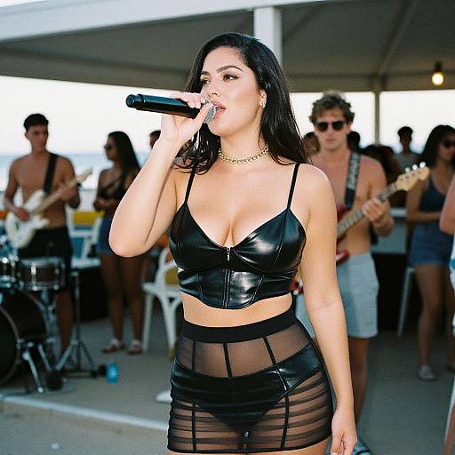 Photograph of a dark-haired woman with medium skin tone, wearing a black leather crop top and sheer black skirt, singing into a microphone at an outdoor