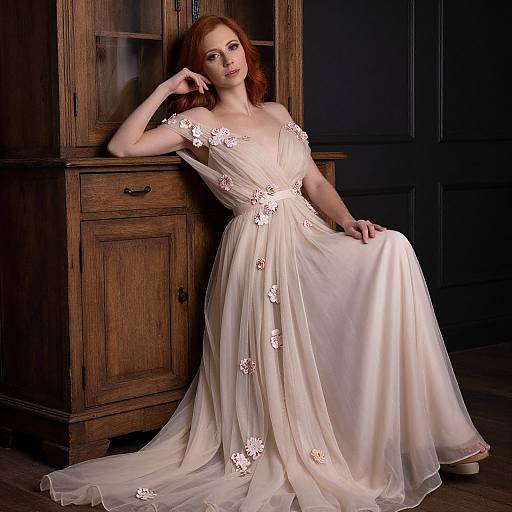 Photograph of a red-haired woman in a white, off-shoulder, floral-embellished gown, leaning against a wooden cabinet in a