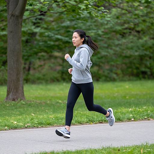 Photograph of an Asian woman jogging on a park path, wearing a gray hoodie, black leggings, and white sneakers, with green trees and grass in