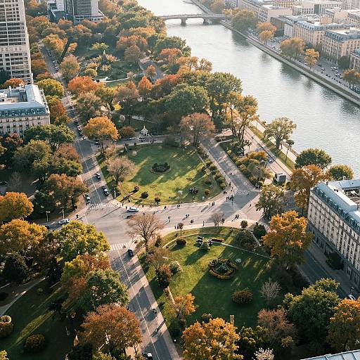 Aerial photograph of a vibrant city park with autumn foliage, people walking, and a river reflecting sunlight, surrounded by buildings.