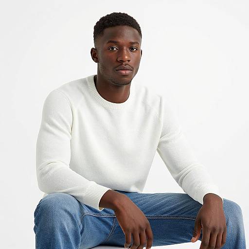 Photograph of a young Black man with short curly hair, wearing a white sweater and blue jeans, sitting against a plain white background, looking directly at