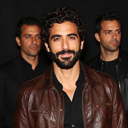 Photograph of three men with Middle Eastern features; central man with dark curly hair, beard, brown leather jacket, black shirt, flanked by two