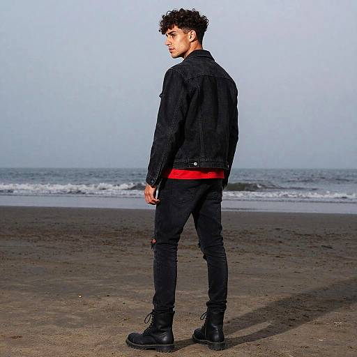 Photograph of a young man with curly brown hair, wearing a black denim jacket, black jeans, and black boots, standing on a beach with a