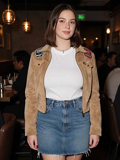 Photograph of a young woman with fair skin and shoulder-length brown hair, wearing a white t-shirt, blue denim skirt, and brown suede jacket with