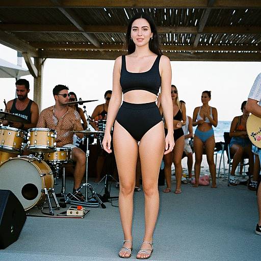 Photograph of a slender, dark-haired woman in black sports bra and high-waisted underwear, standing confidently in front of a band performing under a