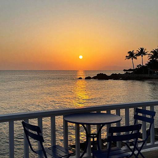 Photograph of a serene sunset over a calm ocean, silhouetted palm trees, and a white railing with a small round table and two chairs