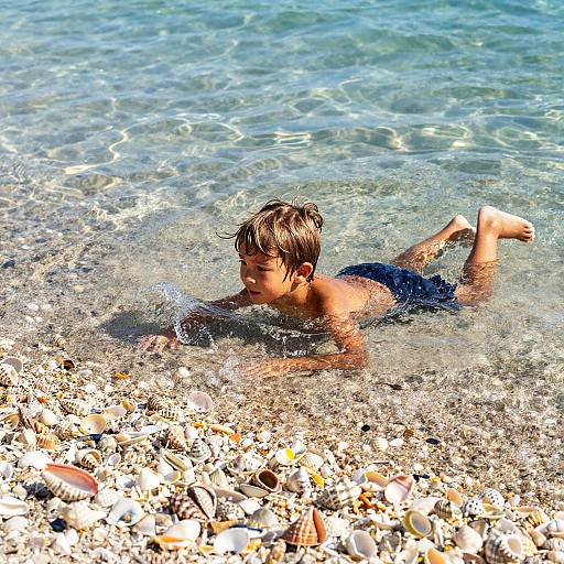 Photograph of a young boy with wet brown hair, wearing dark blue swim trunks, lying on a pebble-covered beach, in clear, shallow