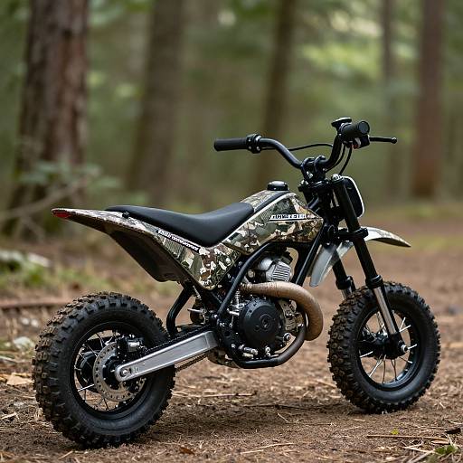 Photograph of a camouflage-patterned dirt bike with black tires and exhaust, parked on a forest dirt path, surrounded by tall trees.