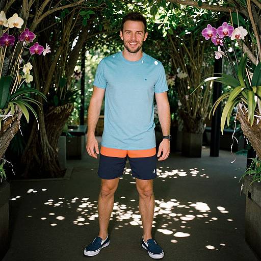 Photograph of a smiling man with short dark hair, light blue shirt, black shorts with orange trim, and blue sneakers, standing in a sunlit