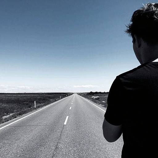 Photograph of a man with dark hair, silhouette in the foreground, standing on a long, empty road with clear blue sky.