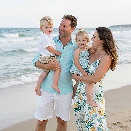 Photograph of a smiling family at the beach: father in light blue shirt and white shorts, mother in floral dress, holding blonde toddler girl, father
