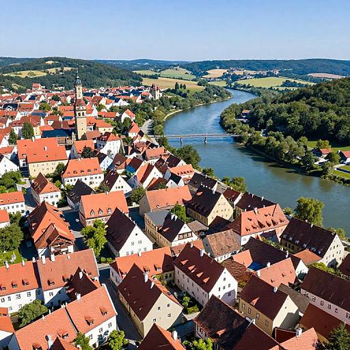 Aerial photograph of a quaint European town with red-roofed houses, a river, and lush greenery, under a clear blue sky.