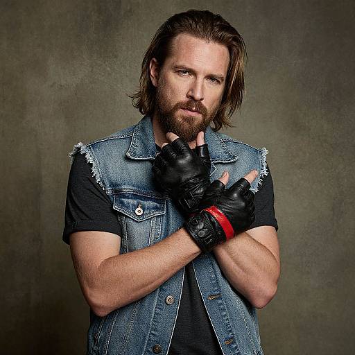 Photograph of a bearded man with long brown hair, wearing a denim vest, black shirt, black leather gloves, and red wristband, against