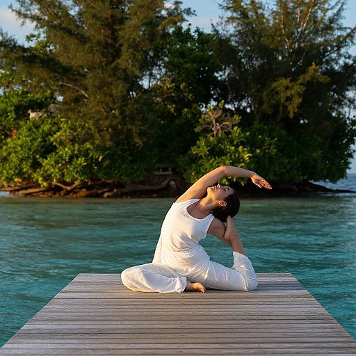 Photograph of a woman in white yoga outfit, performing a yoga pose on a wooden dock, with turquoise water and lush greenery in the background.