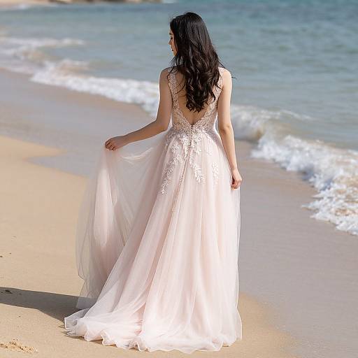 Photograph of a woman with long black hair in a backless, white lace and tulle wedding dress, standing on a sandy beach with gentle waves