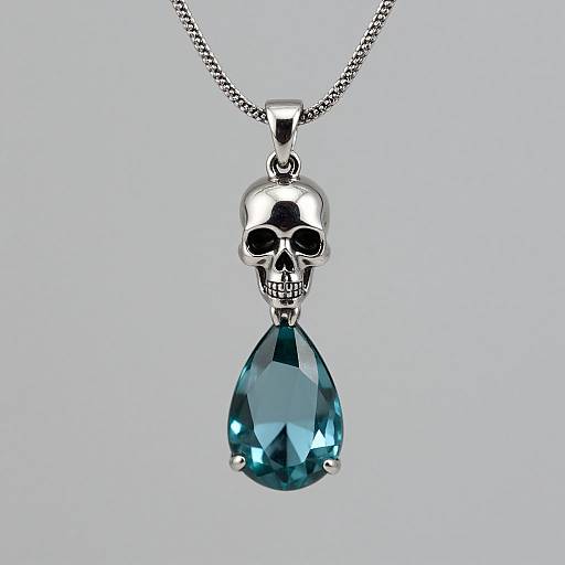 Photograph of a silver skull pendant with a teardrop-shaped blue gemstone, hanging from a delicate chain against a light gray background.