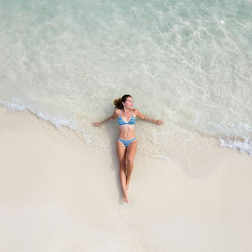 Photograph of a tan-skinned woman in a blue-striped bikini, lying on a sandy beach with clear, shallow ocean water around her.