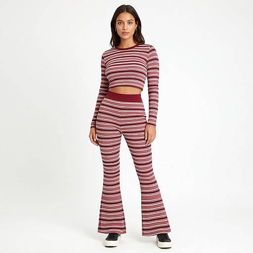 Photograph of a young woman with dark hair, wearing a striped, long-sleeve crop top and matching flared pants, standing against a white