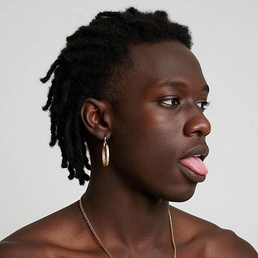 Photograph of a dark-skinned Black woman with short, twisted hair, gold earring, gold necklace, and pink tongue slightly out, against a