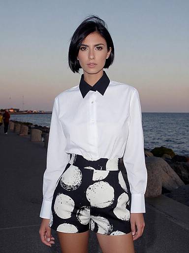 Photograph of a young woman with short black hair, wearing a white long-sleeve shirt with black collar and black high-waisted shorts with