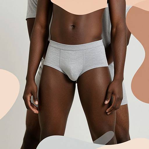 Photograph of a dark-skinned man standing, wearing light gray textured boxer briefs, with abstract peach and white shapes in background.