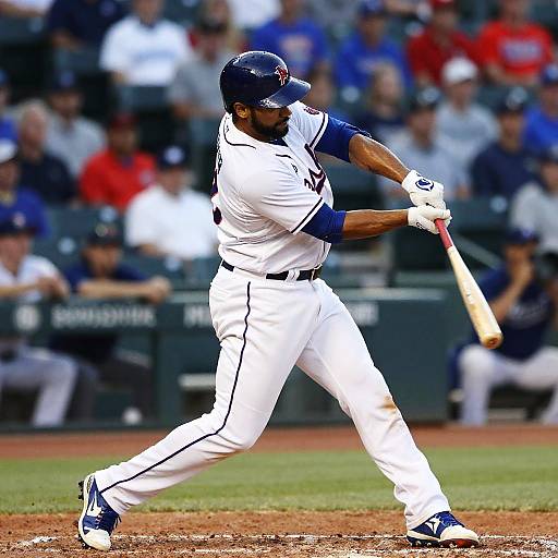 Photograph of a muscular Black baseball player in white uniform, mid-swing with a wooden bat, on a dirt field, with blurred spectators in the