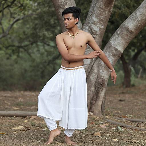 Shirtless young man with dark skin and black hair, wearing white dhoti and gold jewelry, dances in a forest clearing.