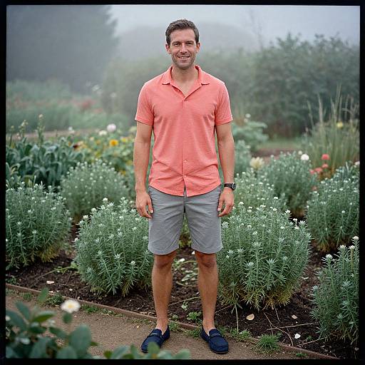 Photograph of a smiling man in a coral polo shirt, gray shorts, and navy shoes, standing in a misty garden with greenery and white