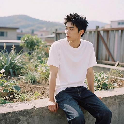 Photograph of an Asian young man with spiky black hair, wearing a white t-shirt and dark jeans, sitting on a concrete garden wall, looking